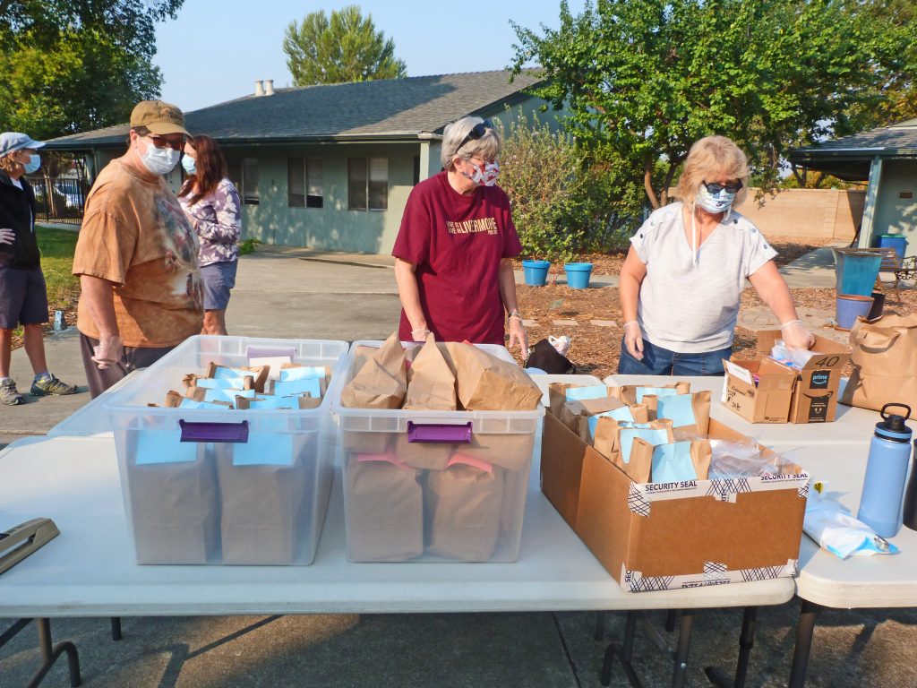 Three female congregation members stand behind a folding table while organizing brown paper bags for the homeless