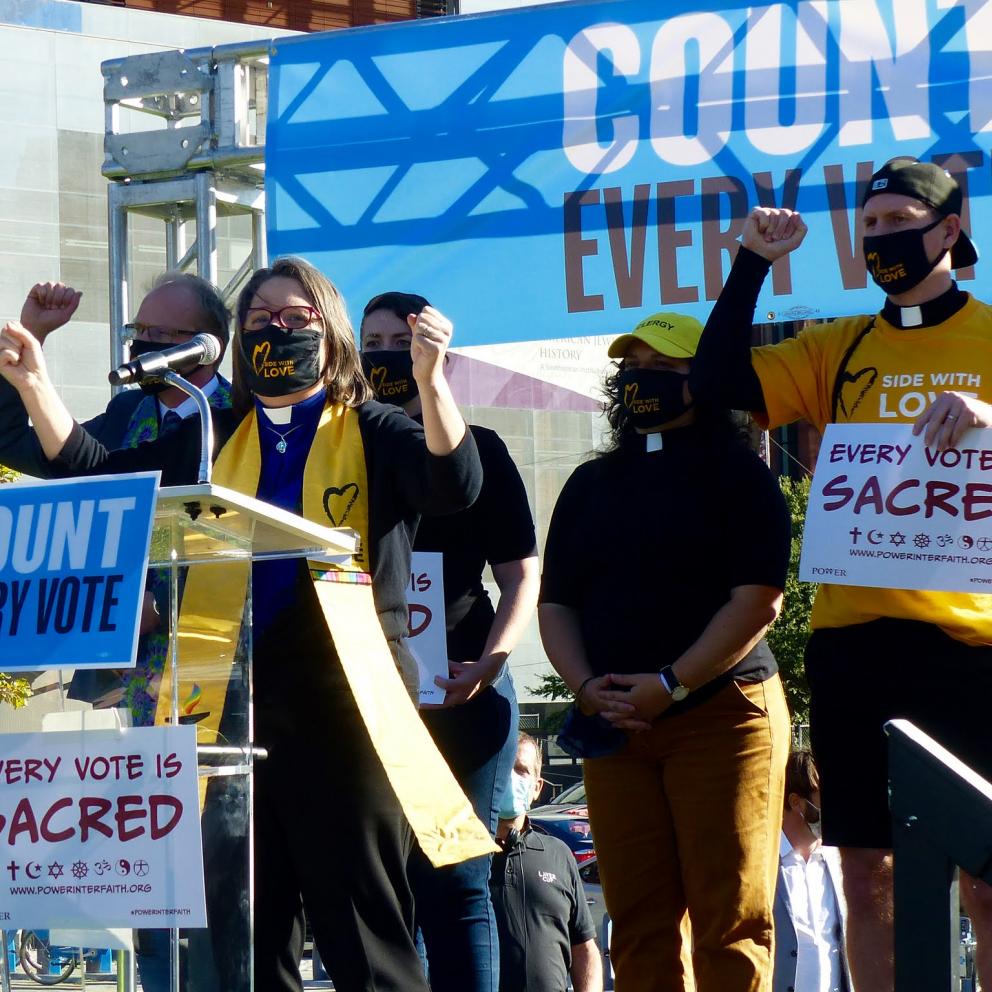 Five people wearing yellow and standing on a stage, one person is behind a lectern, a banner with "Count Every Vote" is behind the people