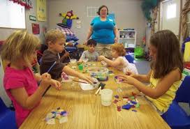 Five children doing crafts at a table while being directed by an adult